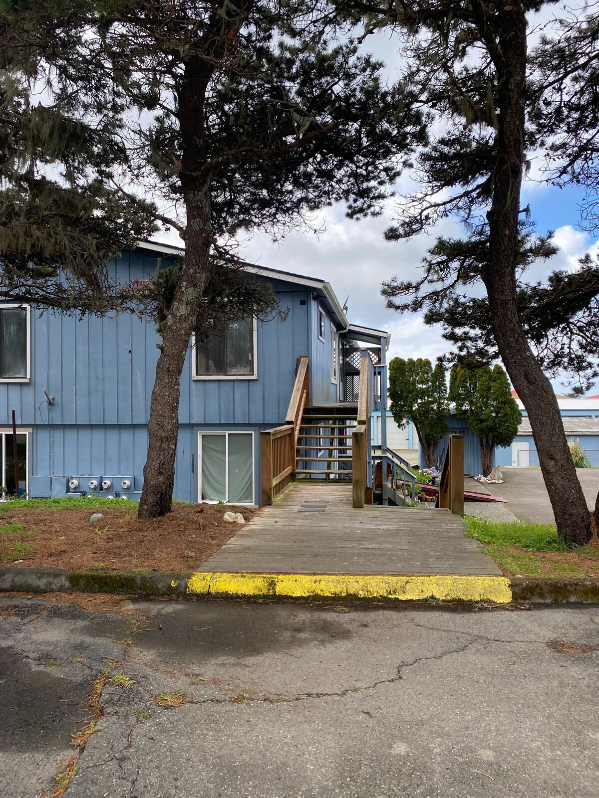 Blue two-story apartment building framed by coastal pines at the Coastal Apartments & Storage property